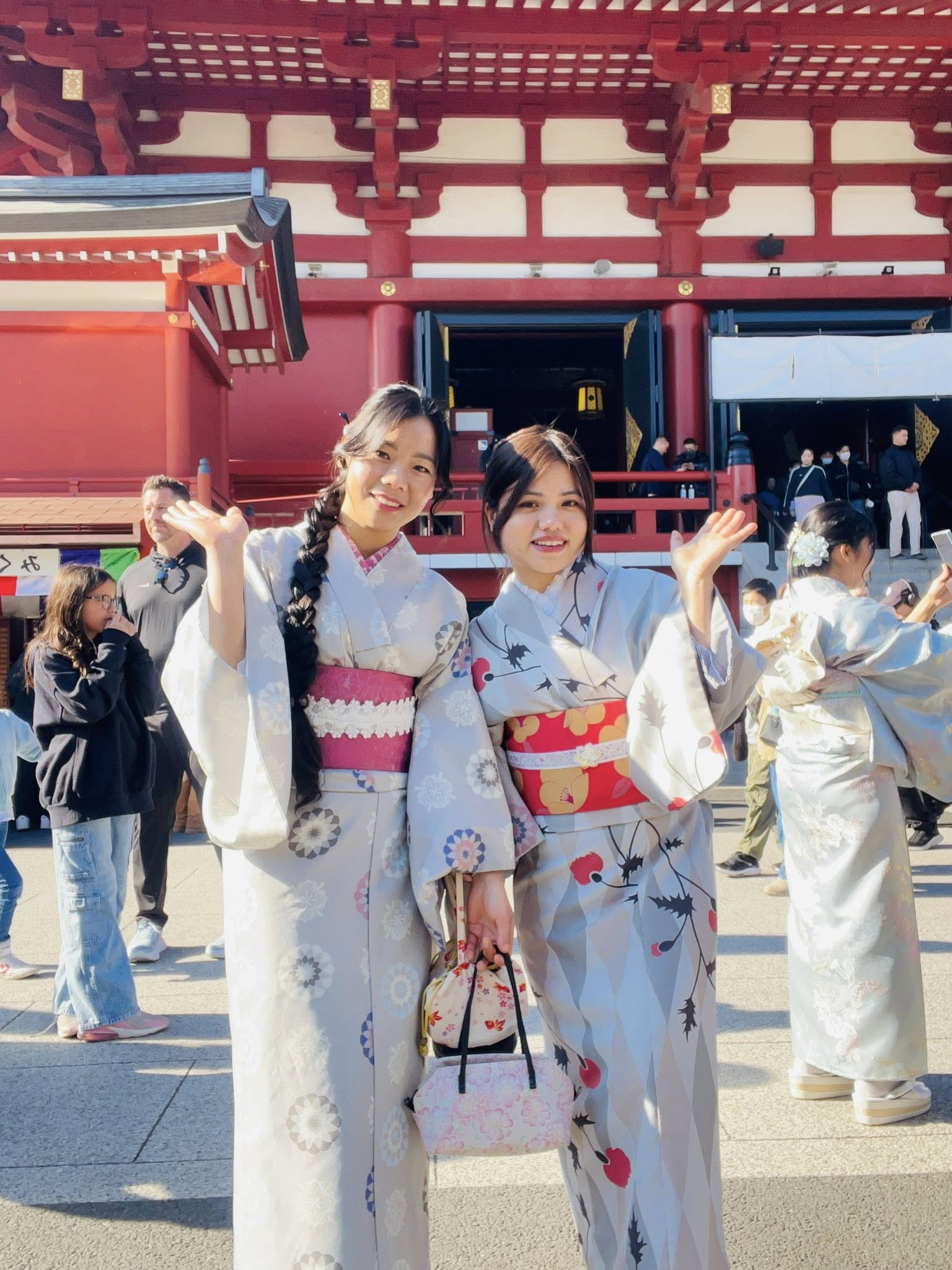 Two friends in traditional kimono waving, in front of a red Japanese temple